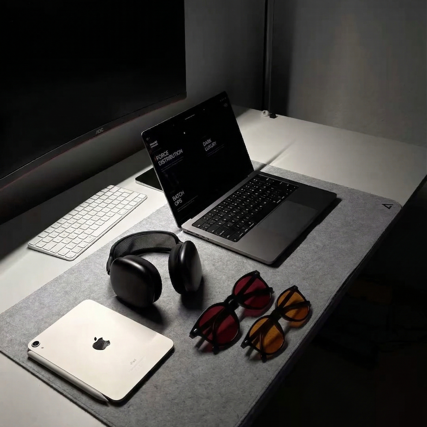 Dark office desk with laptop, headphones, tablet, and both red night and orange day blue light glasses on a gray mat.