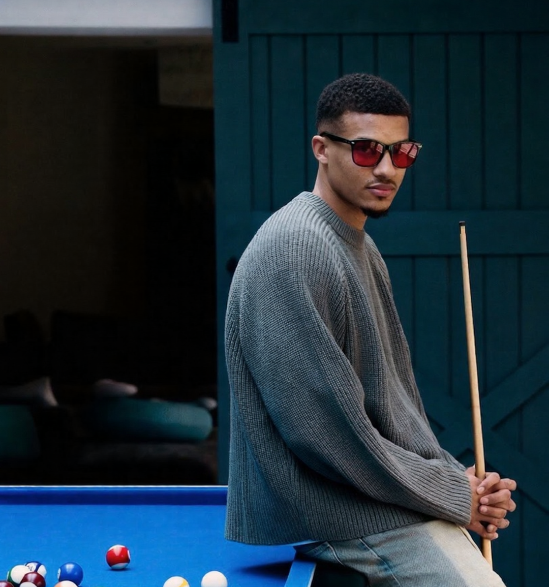 Man in a gray sweater holding a pool cue next to a pool table with pool balls.