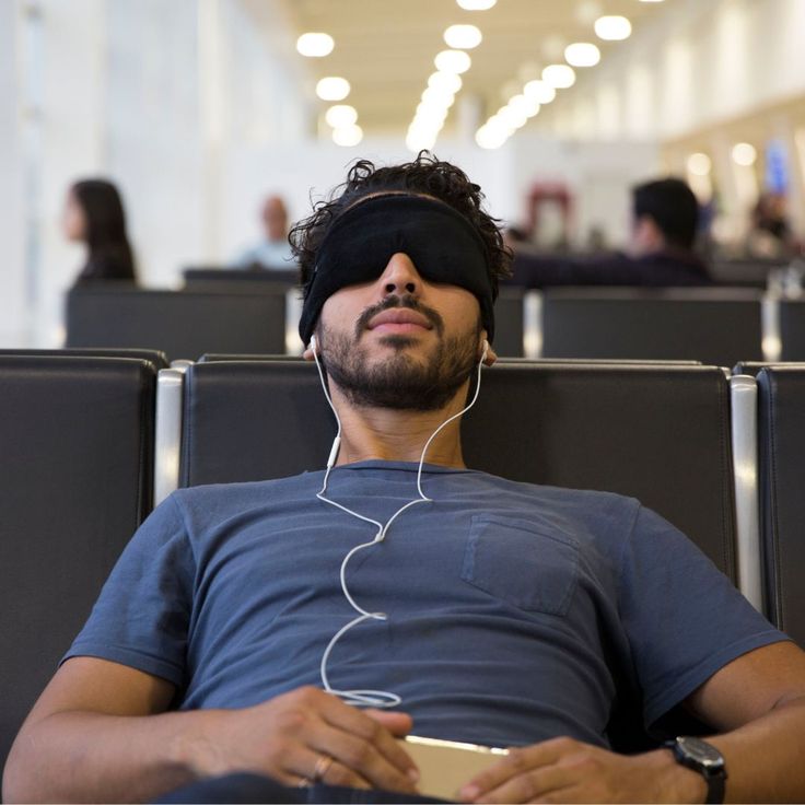 A man reclining in an airport or train station seating area, wearing a black sleep mask and white wired earbuds, with a smartphone in his hands.